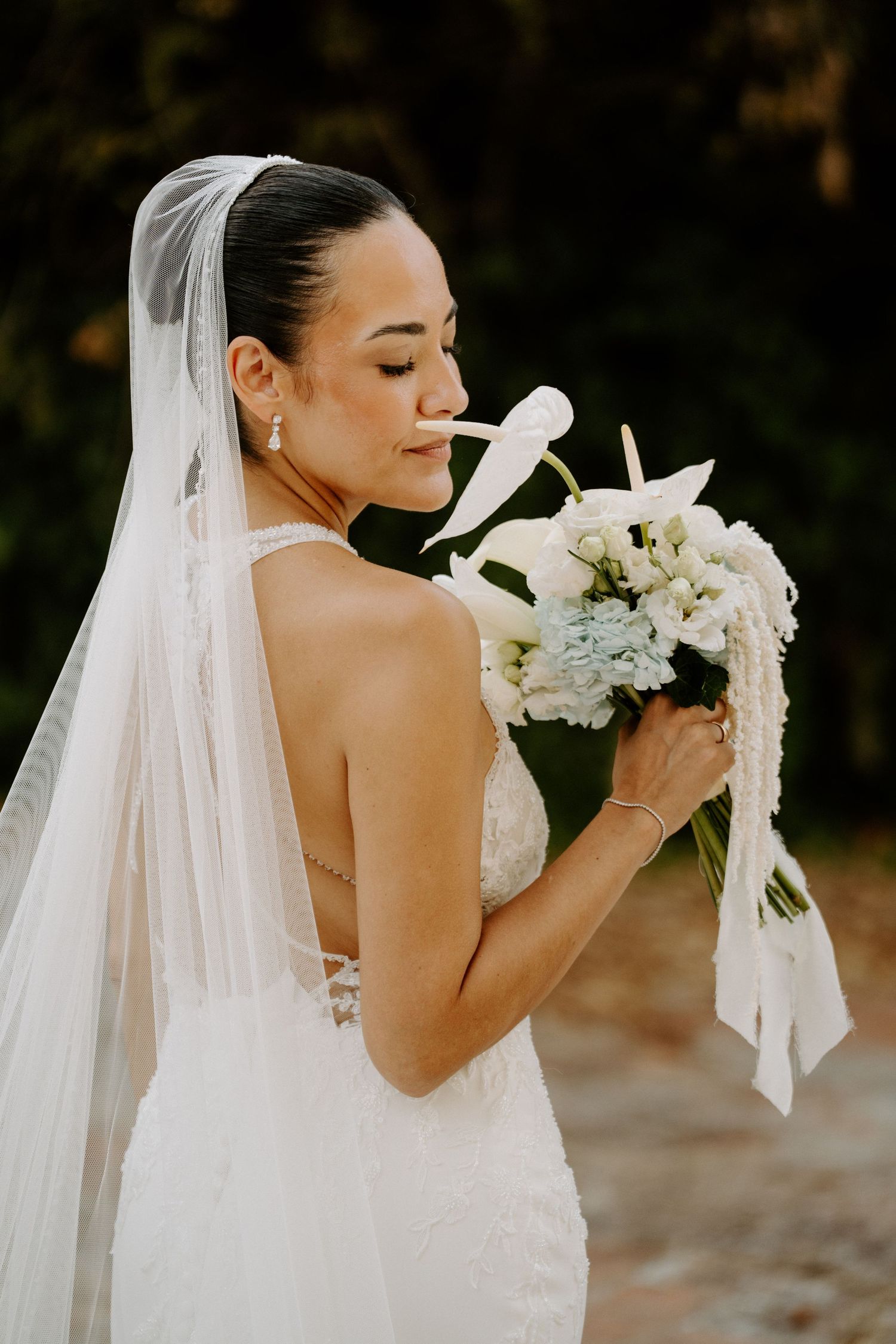 Elegant bride in a halter gown and veil smells her white and blue bridal bouquet in a serene outdoor setting.