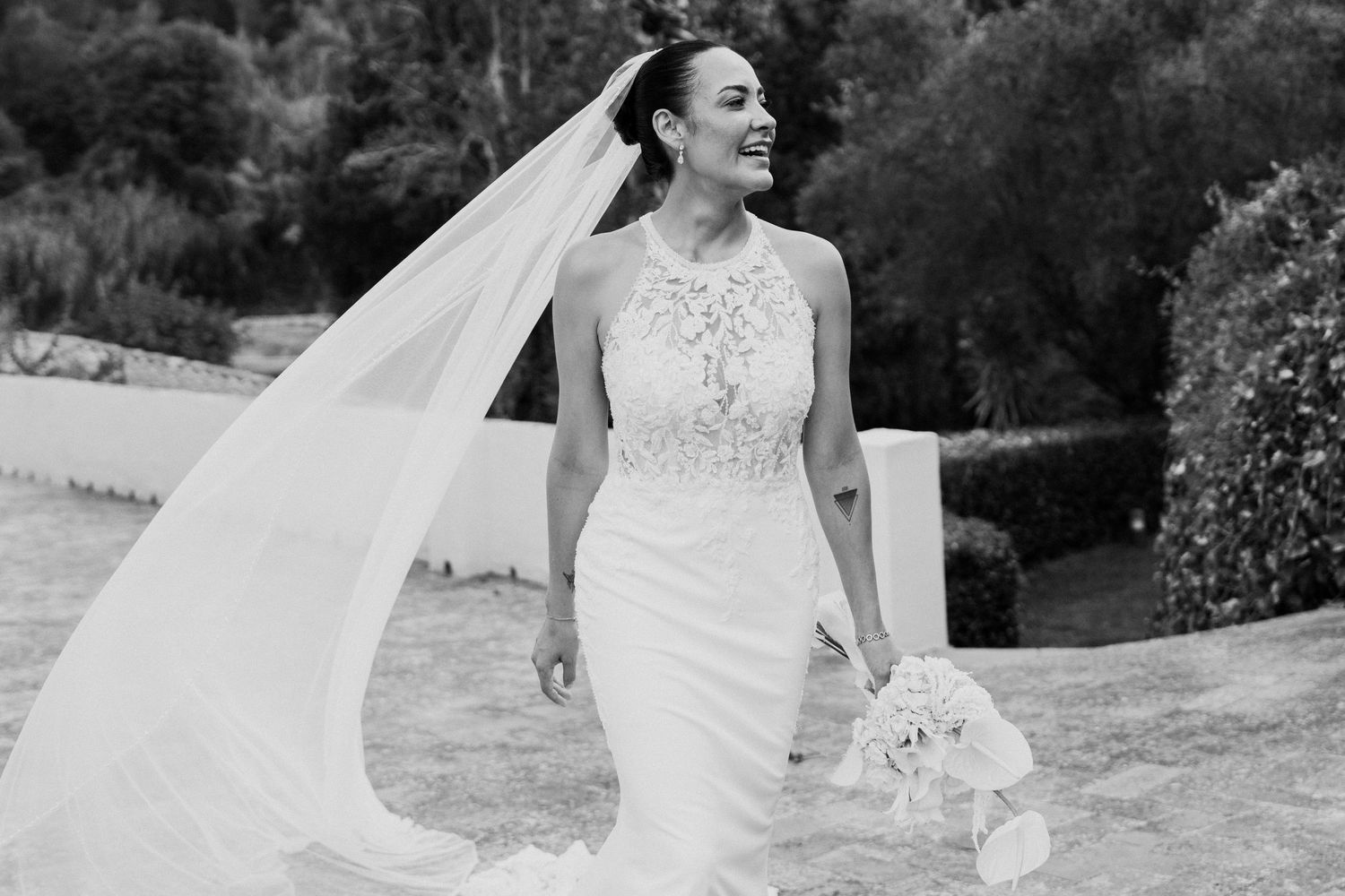 Black and white photo of smiling bride in halter wedding gown with flowing veil holding bouquet.