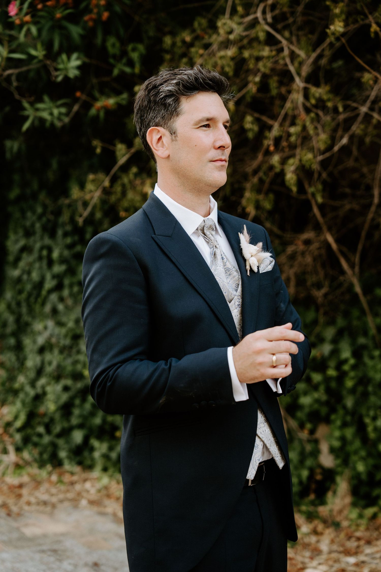 Dapper groom in a navy suit with floral boutonniere and paisley tie stands confidently in an outdoor garden setting.