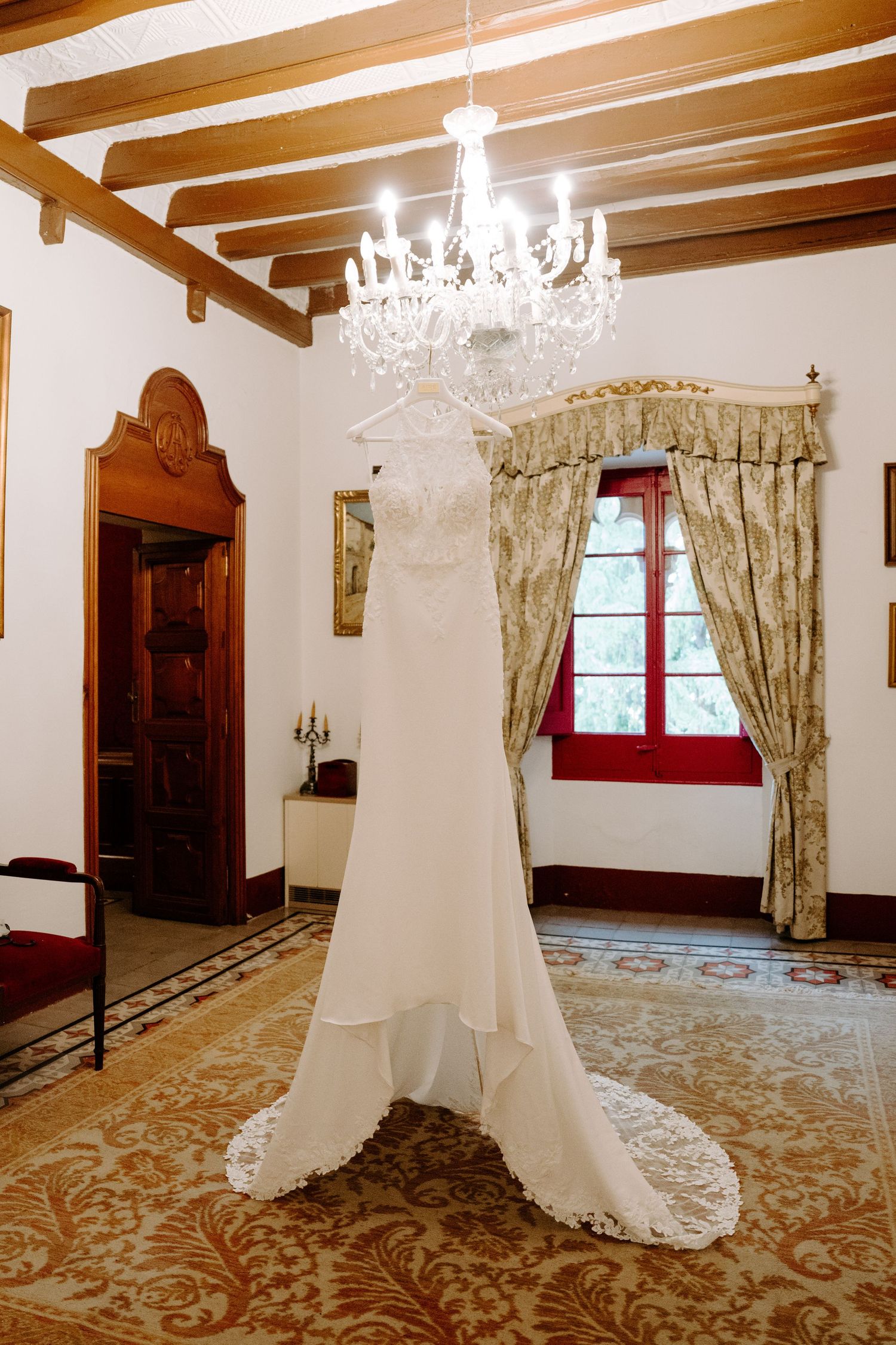 Elegant white wedding dress with lace train hanging from crystal chandelier in a rustic venue with wooden beam ceiling.