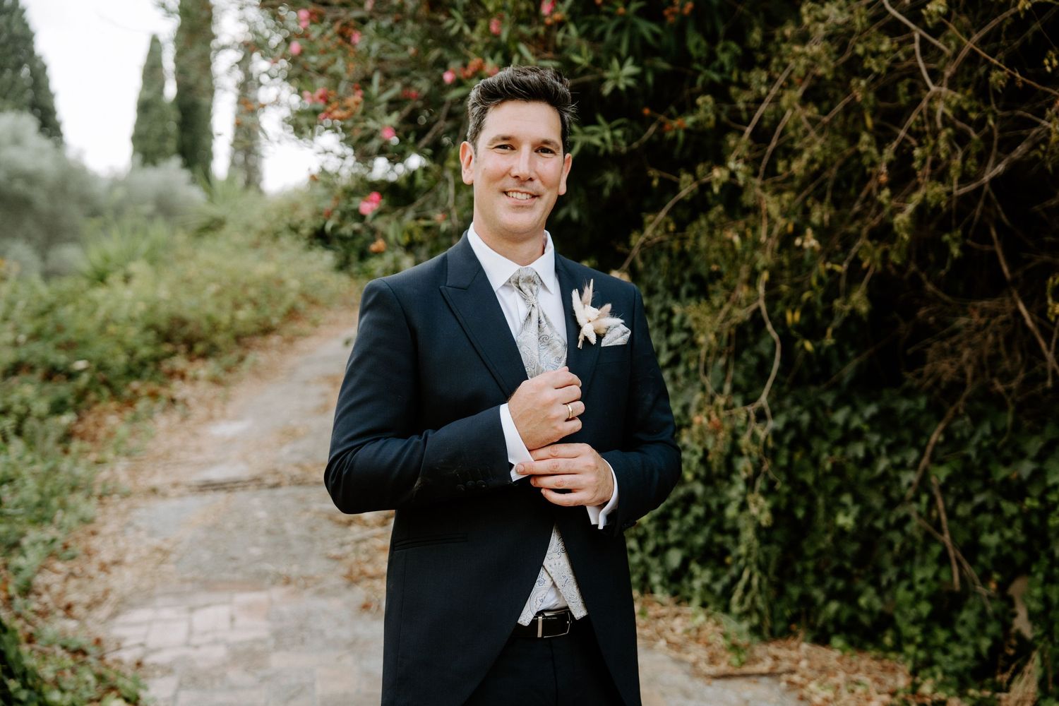 Dapper groom in navy suit with floral boutonniere standing on garden pathway among lush greenery.