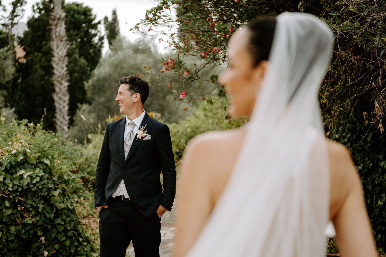 Groom in black suit gazes lovingly at bride in white veil among lush garden greenery.