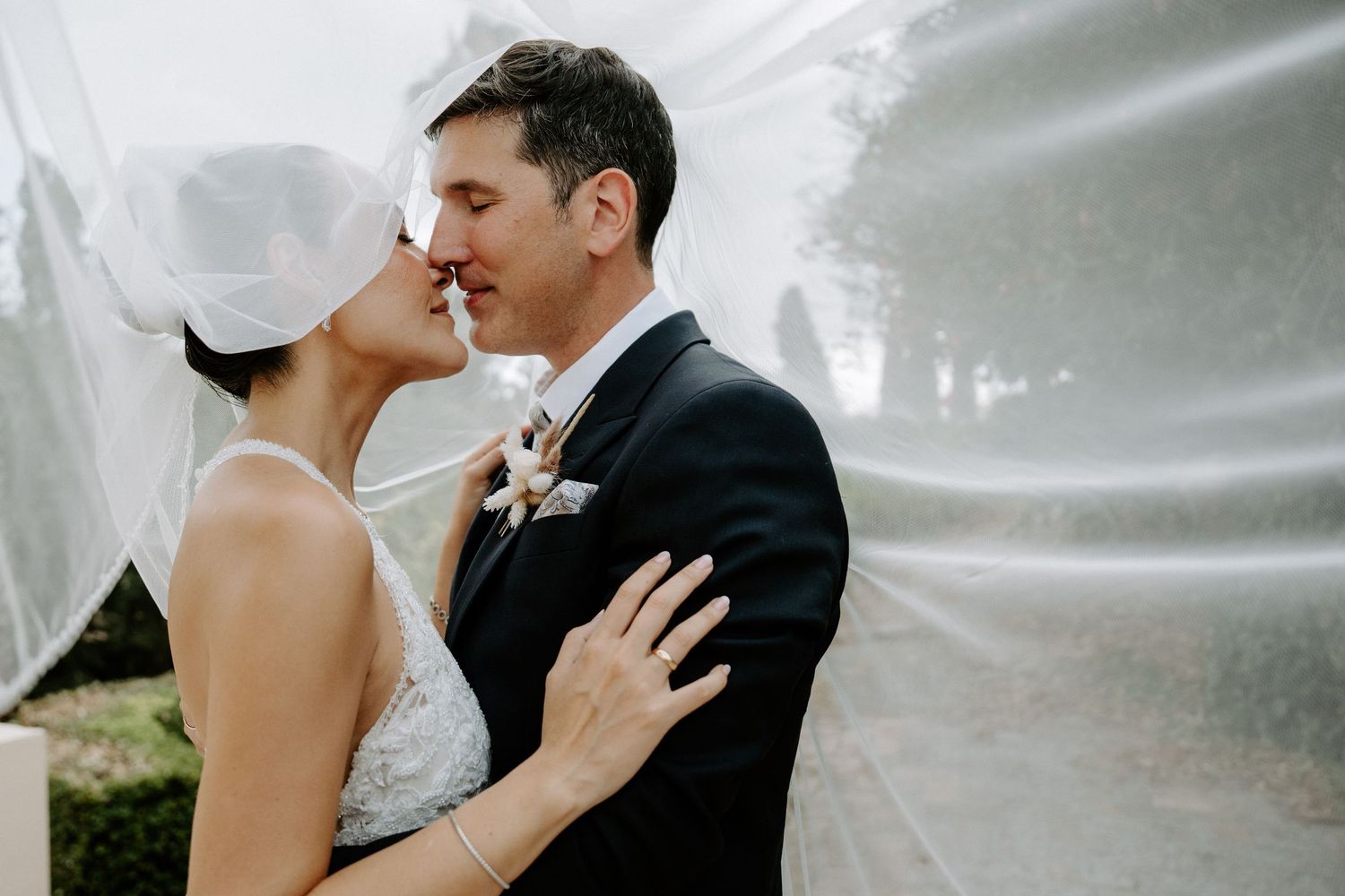 Romantic couple nearly kissing under flowing bridal veil in soft natural outdoor lighting.