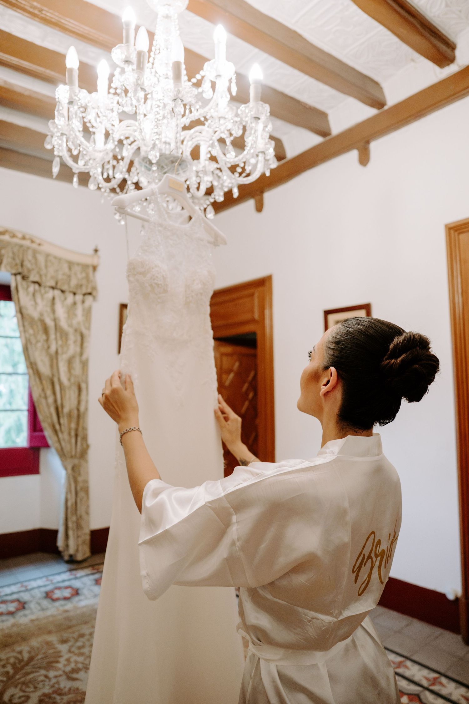 Bride in white satin robe adjusting her lace wedding dress hanging from an elegant crystal chandelier.