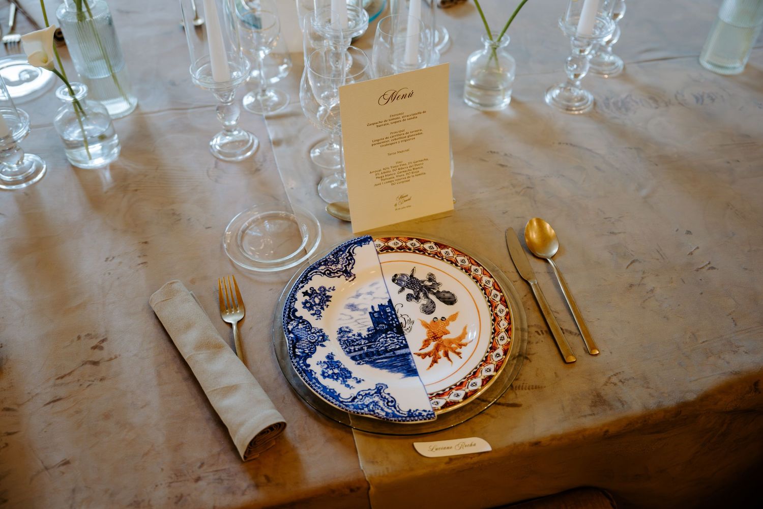 Elegant wedding place setting with blue and white chinoiserie plate, gold cutlery, crystal glasses, and a menu card on linen.