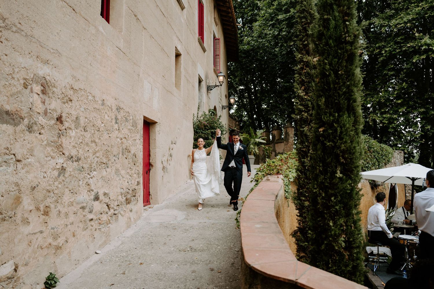 Bride and groom walking along stone pathway beside rustic building with cypress trees at wedding venue.