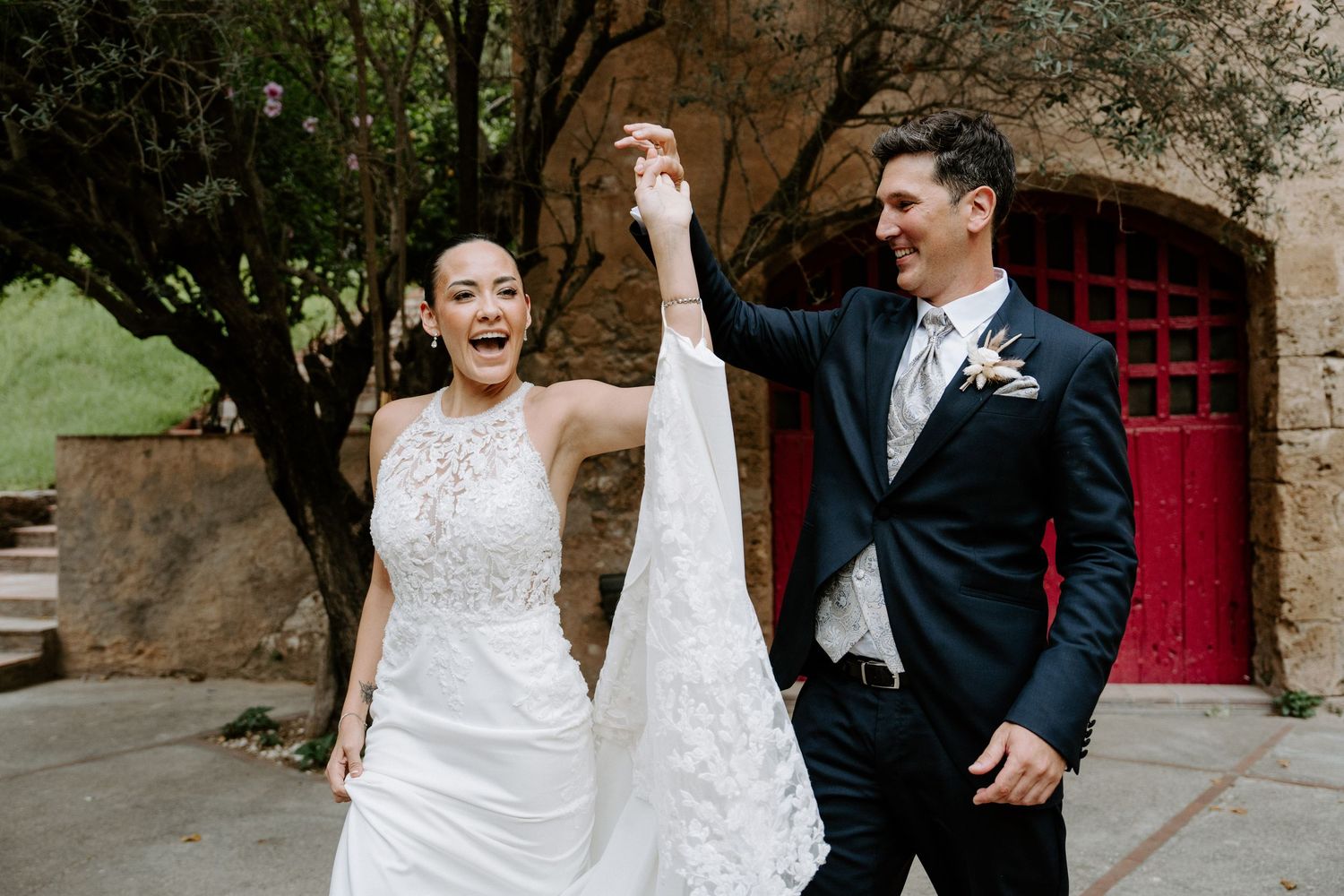 Joyful bride and groom walking outdoors, bride in halter wedding gown with lace veil, groom in dark suit, red doors behind.