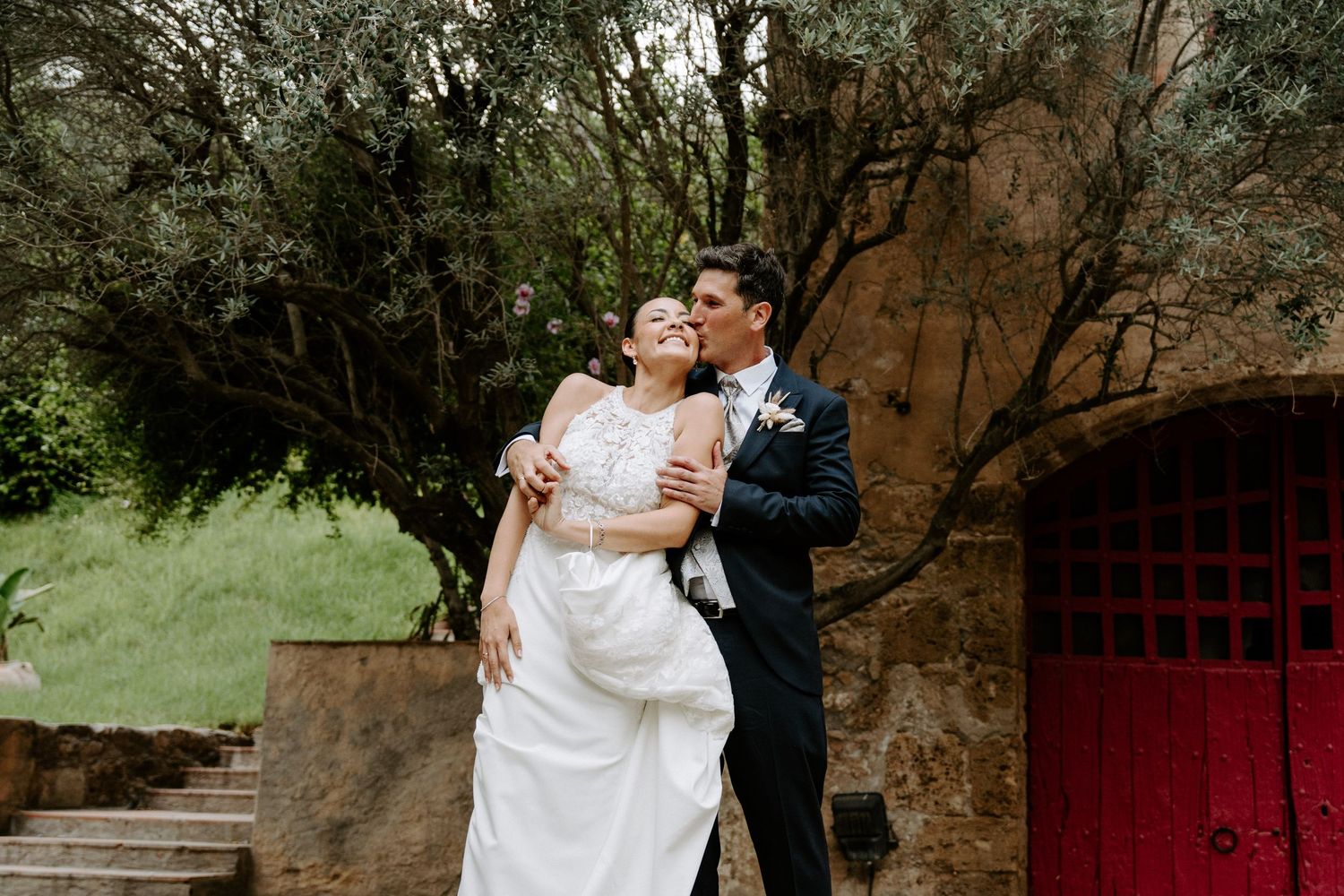 Groom kisses bride on cheek as she laughs joyfully outdoors near red door and olive trees.