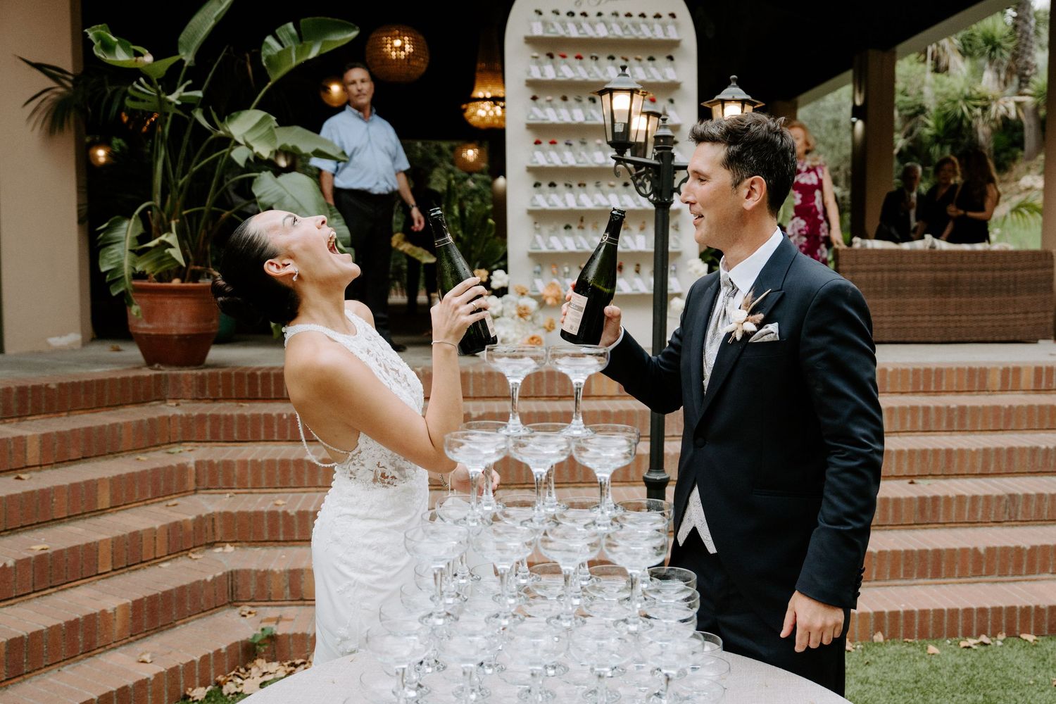 Bride and groom pouring champagne tower at outdoor wedding reception, laughing and celebrating together.