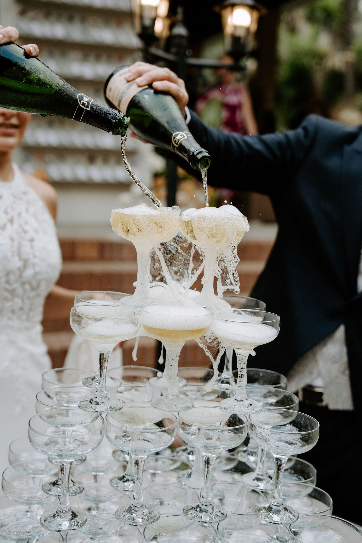 Champagne tower overflowing as bride and groom pour bottles over stacked coupe glasses at reception.
