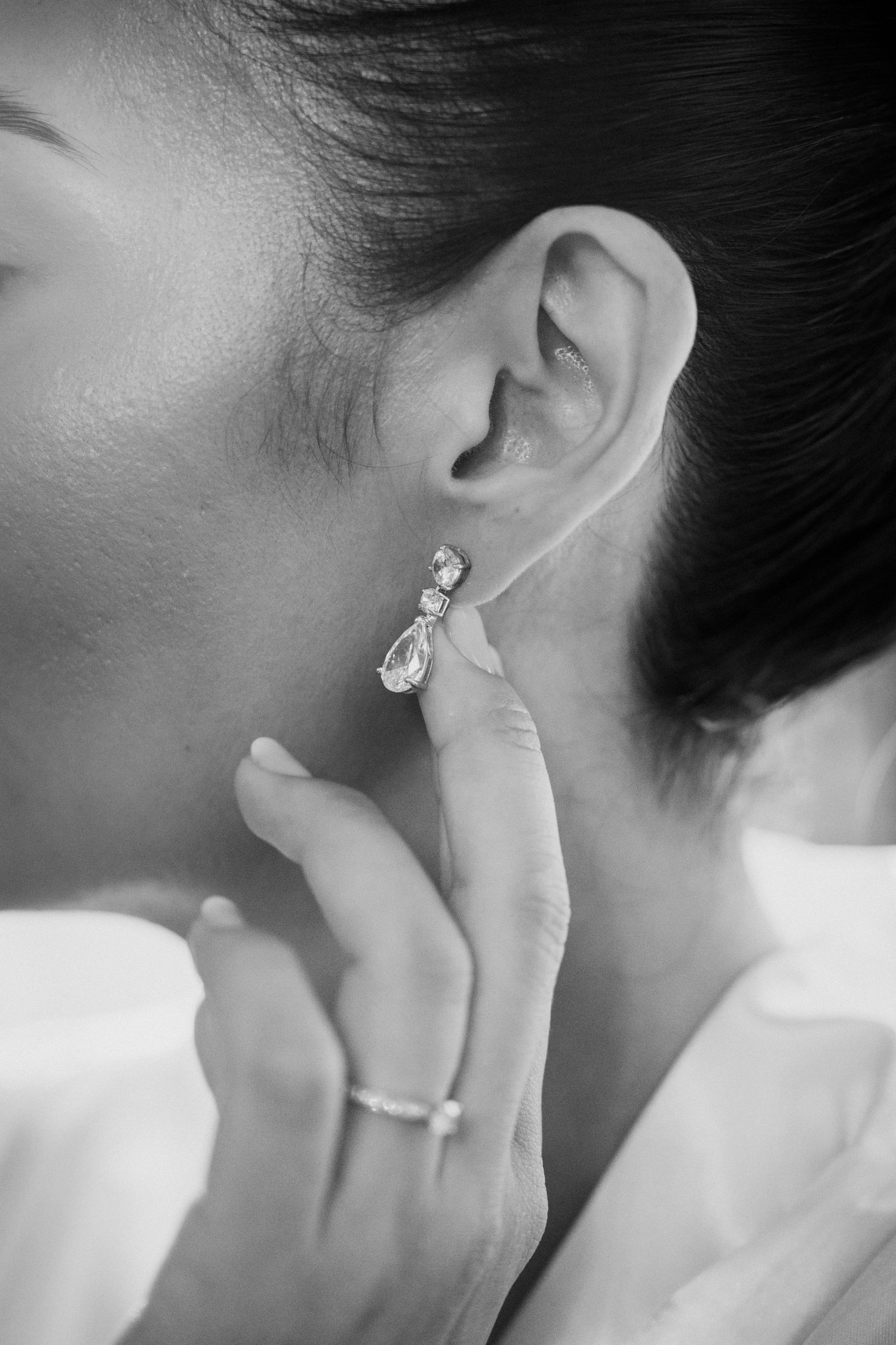 Black and white close-up of bride putting on a crystal drop earring with a diamond ring on her finger.