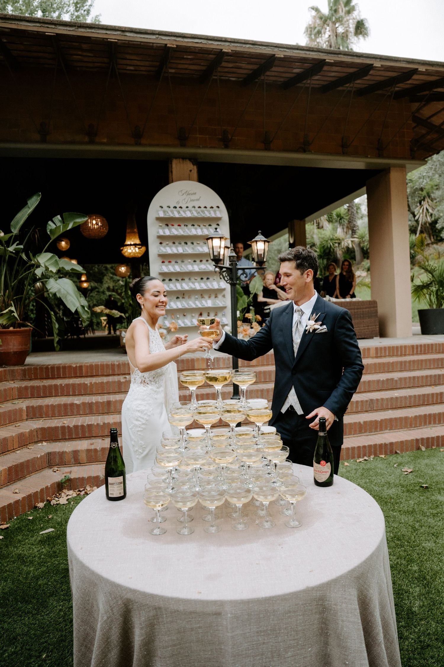 Newlyweds pour champagne tower together outdoors at elegant garden wedding reception celebration.