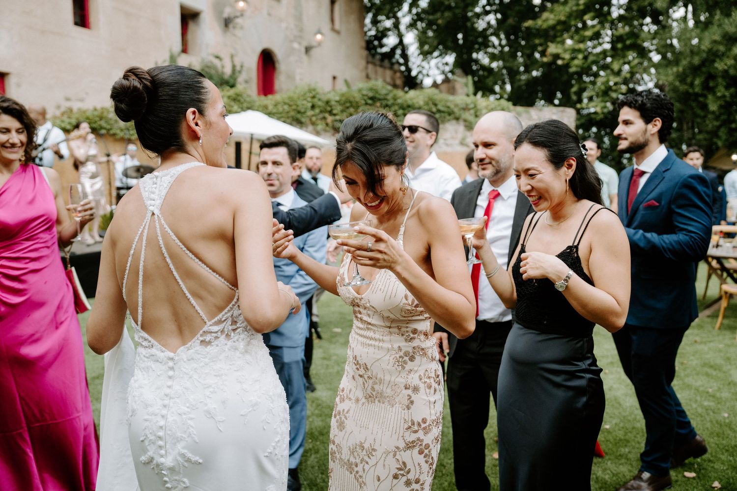 Guests laughing and celebrating at an outdoor wedding reception, with bride in backless white gown.