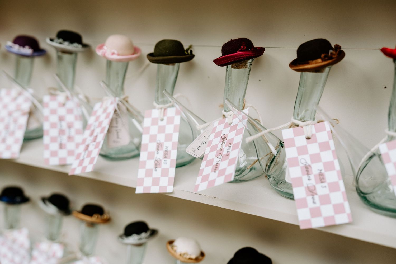 Decorative mini hats on glass vases with pink checkered gift tags displayed on white shelves as wedding favors.
