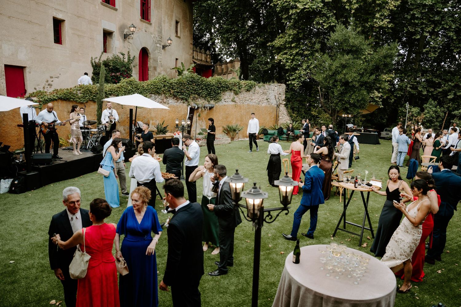Wedding guests mingle on a sunlit lawn during an outdoor cocktail hour at a rustic European venue.