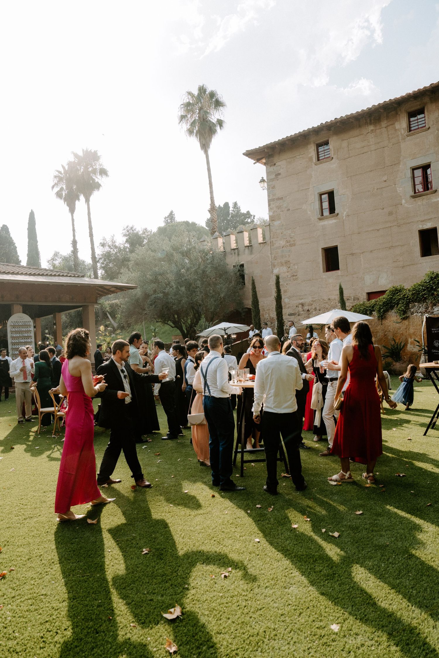 Elegantly dressed wedding guests socializing outdoors at historic stone manor with palm trees in golden light.