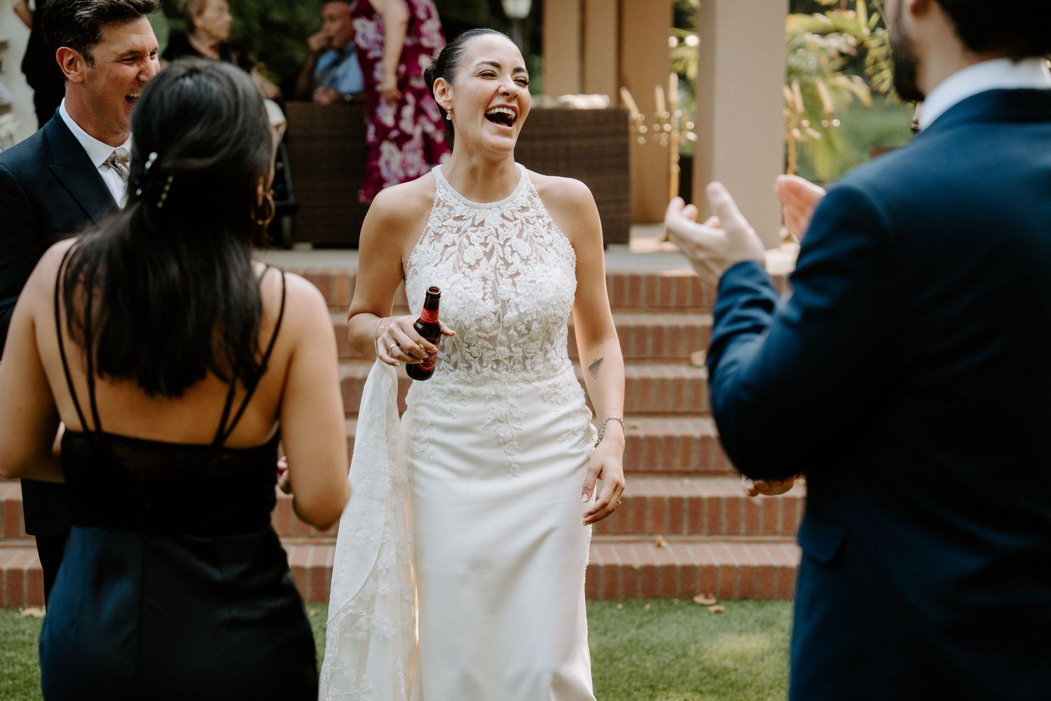 Bride in white halter gown laughing joyfully with guests at an outdoor wedding reception.