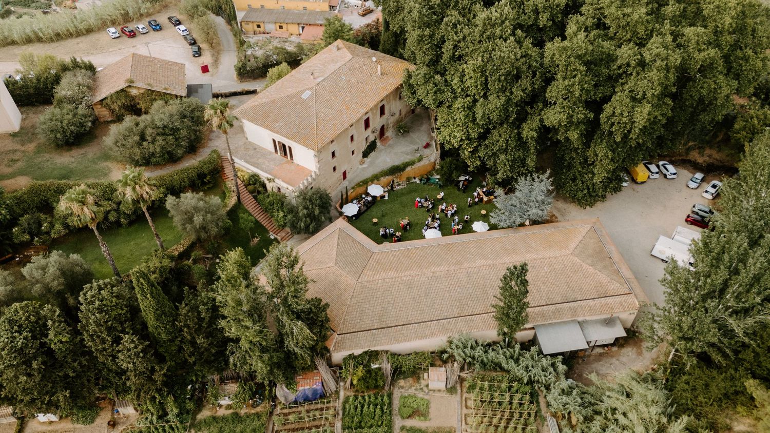 Aerial view of a rustic stone farmhouse surrounded by lush green trees with outdoor seating and parked cars nearby.