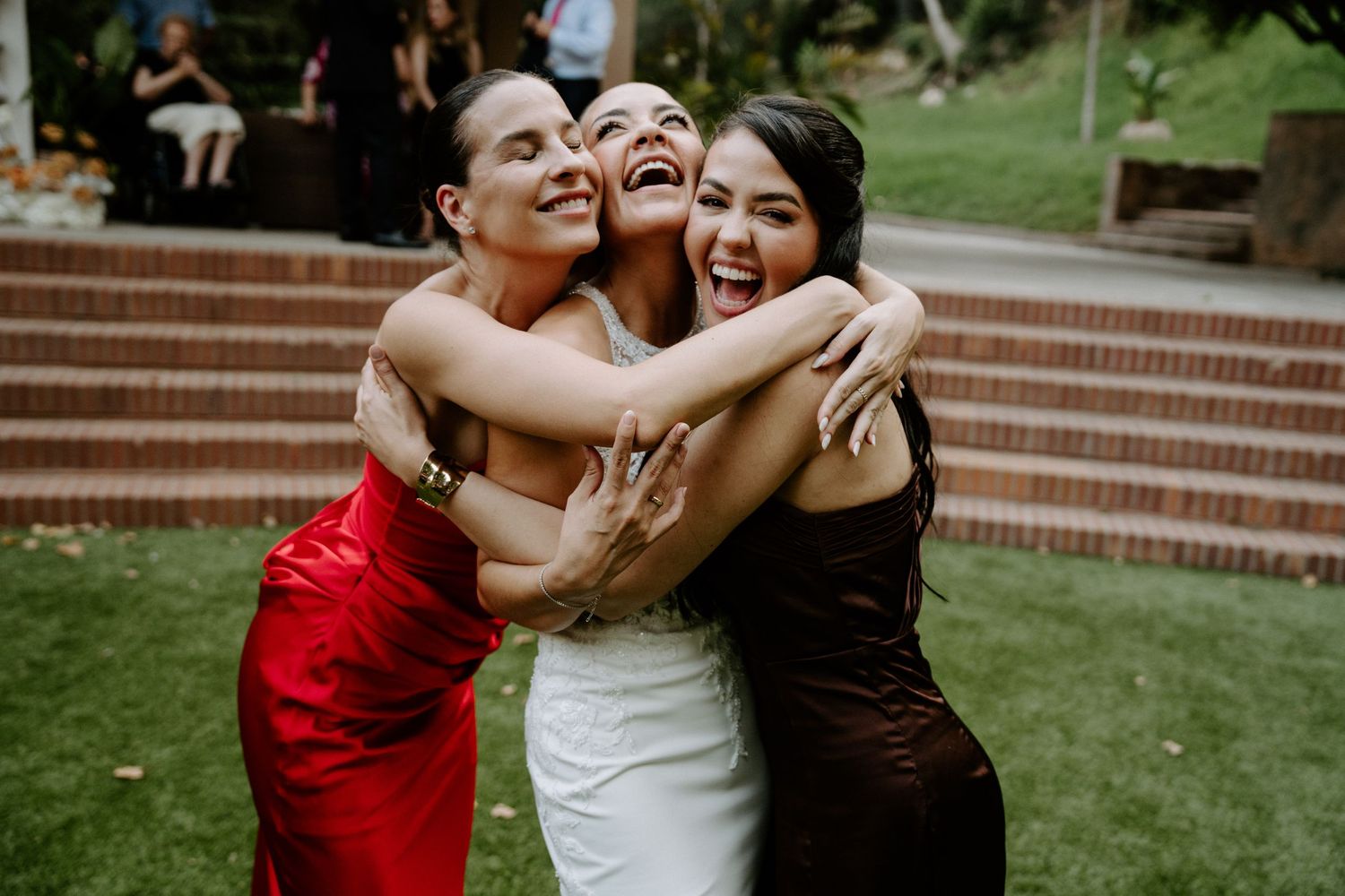 Three women in elegant dresses joyfully embrace outdoors at a wedding celebration.