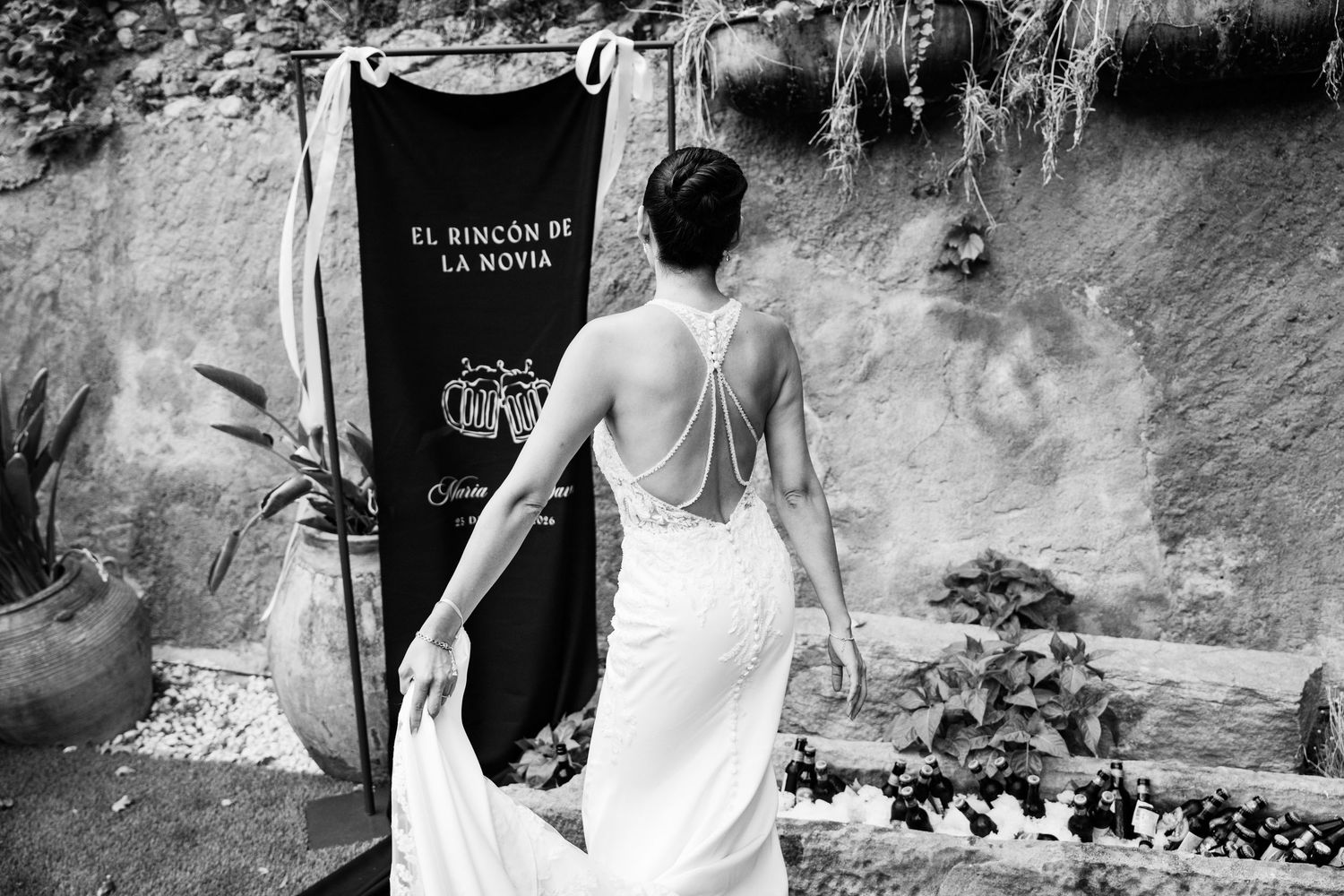 Bride in a backless lace wedding gown standing before a sign at El Rincon de la Novia in black and white photo.
