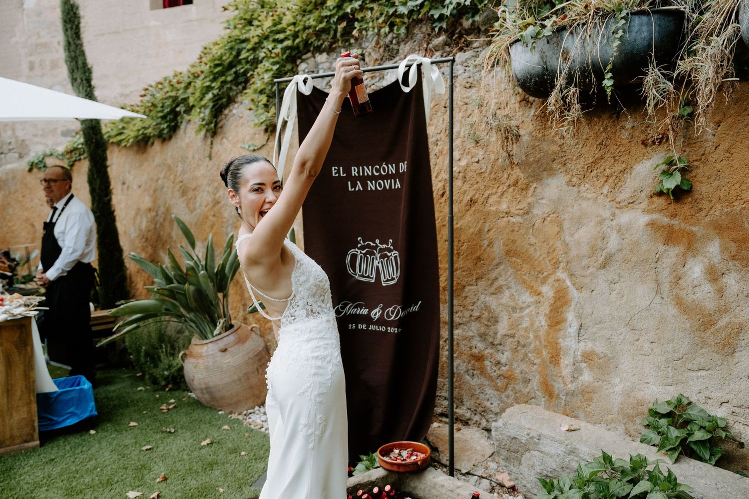 Bride in white gown adjusting a decorative wedding sign on a rustic stone wall outdoors.