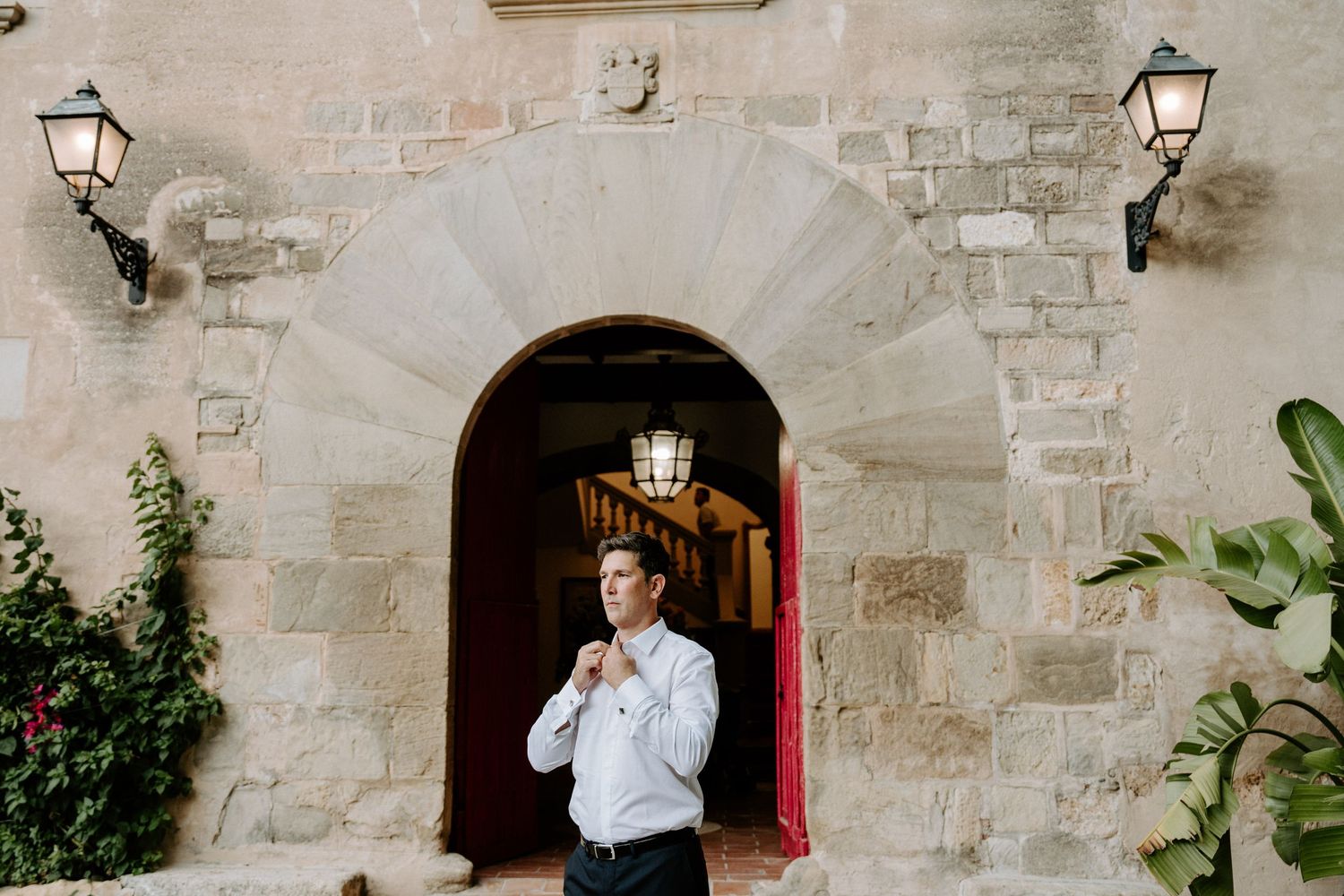 Groom adjusts his tie under a stone archway with red doors and lanterns at a wedding venue.