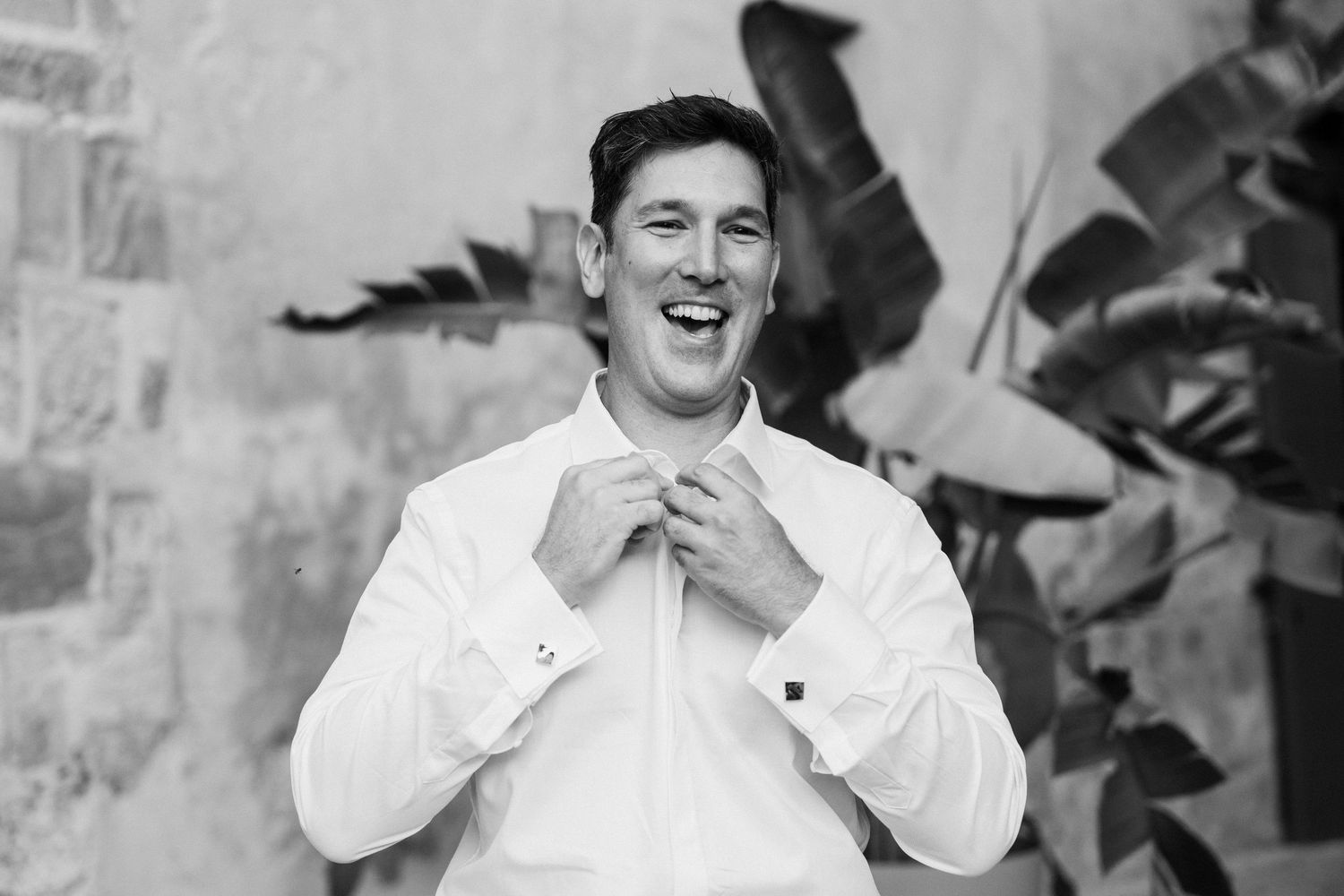Smiling groom in white dress shirt adjusting his collar in a black and white wedding photo.