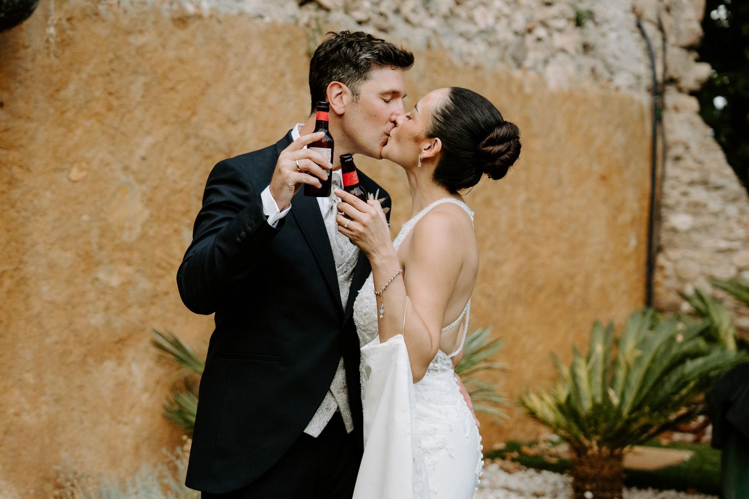 Bride and groom share a romantic kiss while holding drinks at an outdoor wedding celebration near a stone wall.