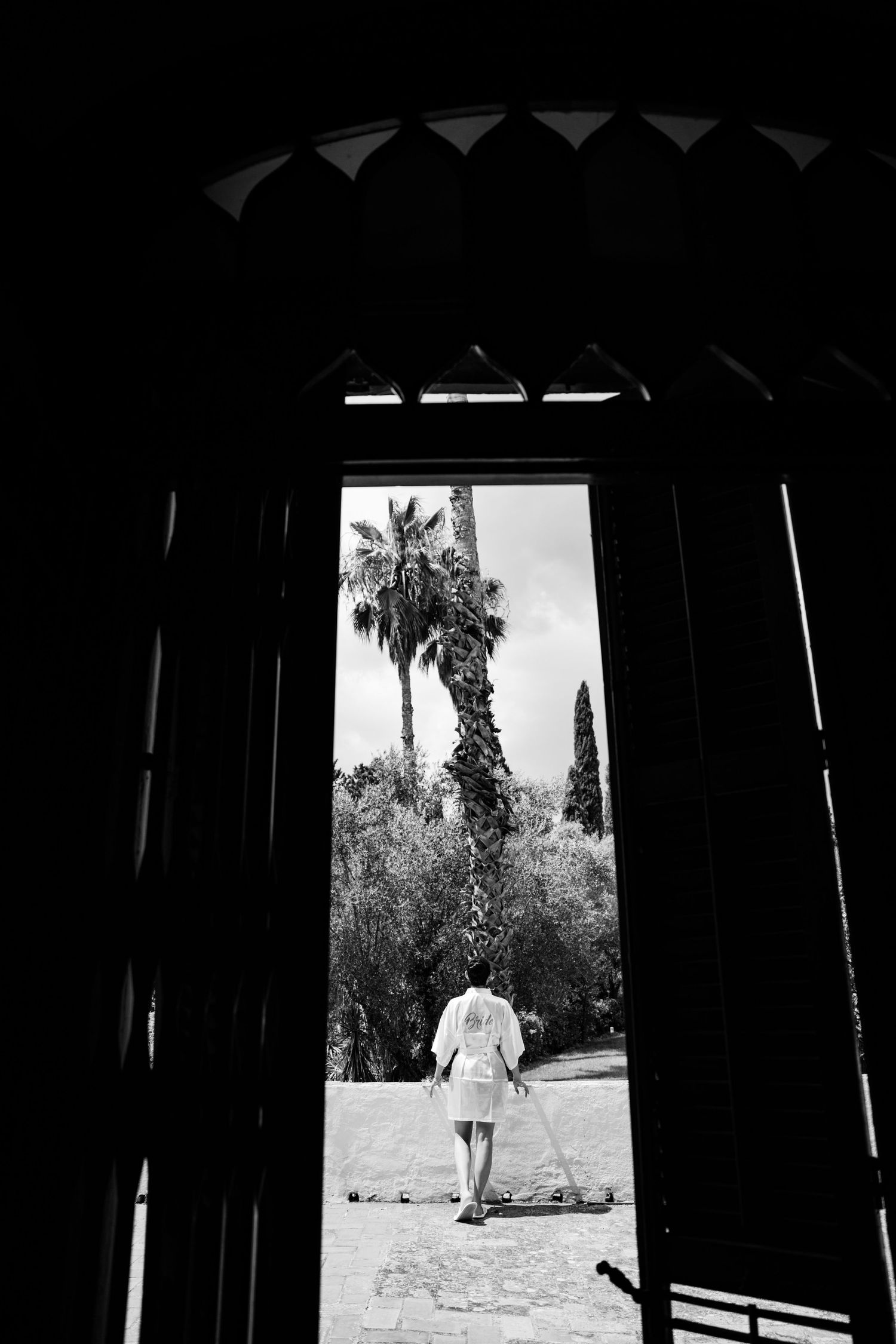 Black and white photo of a bride walking through ornate doors toward palm trees in bright sunlight.