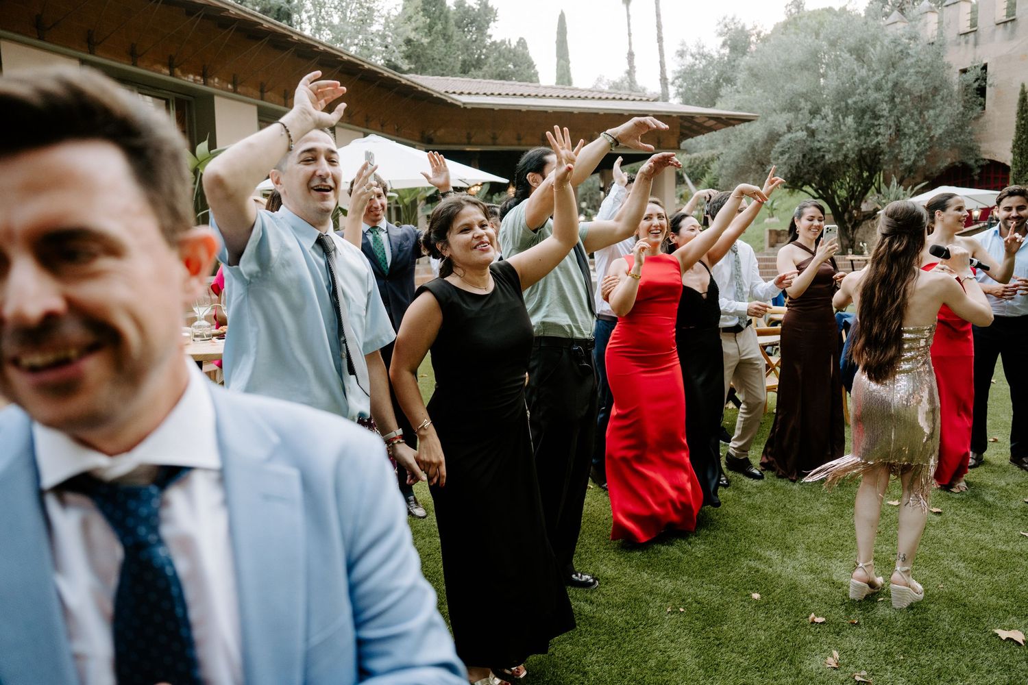 Excited wedding guests raising hands and dancing together during an outdoor reception party.