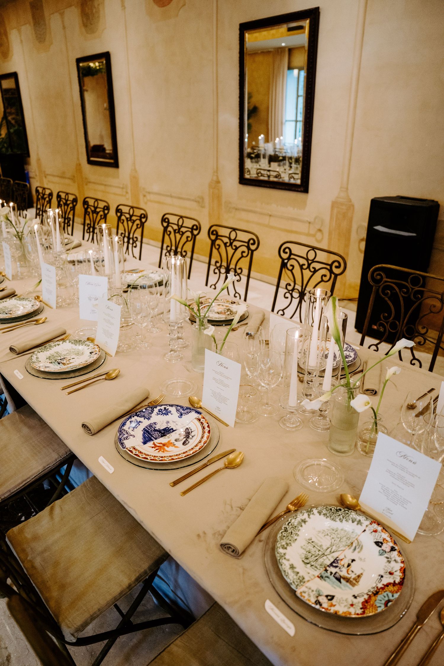 Elegantly set long reception table with ornate plates, gold cutlery, crystal glasses, and candles in a classic venue.