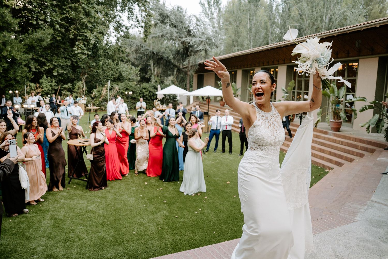 Joyful bride in white gown tossing bouquet to excited wedding guests gathered on a lush green lawn outdoors.