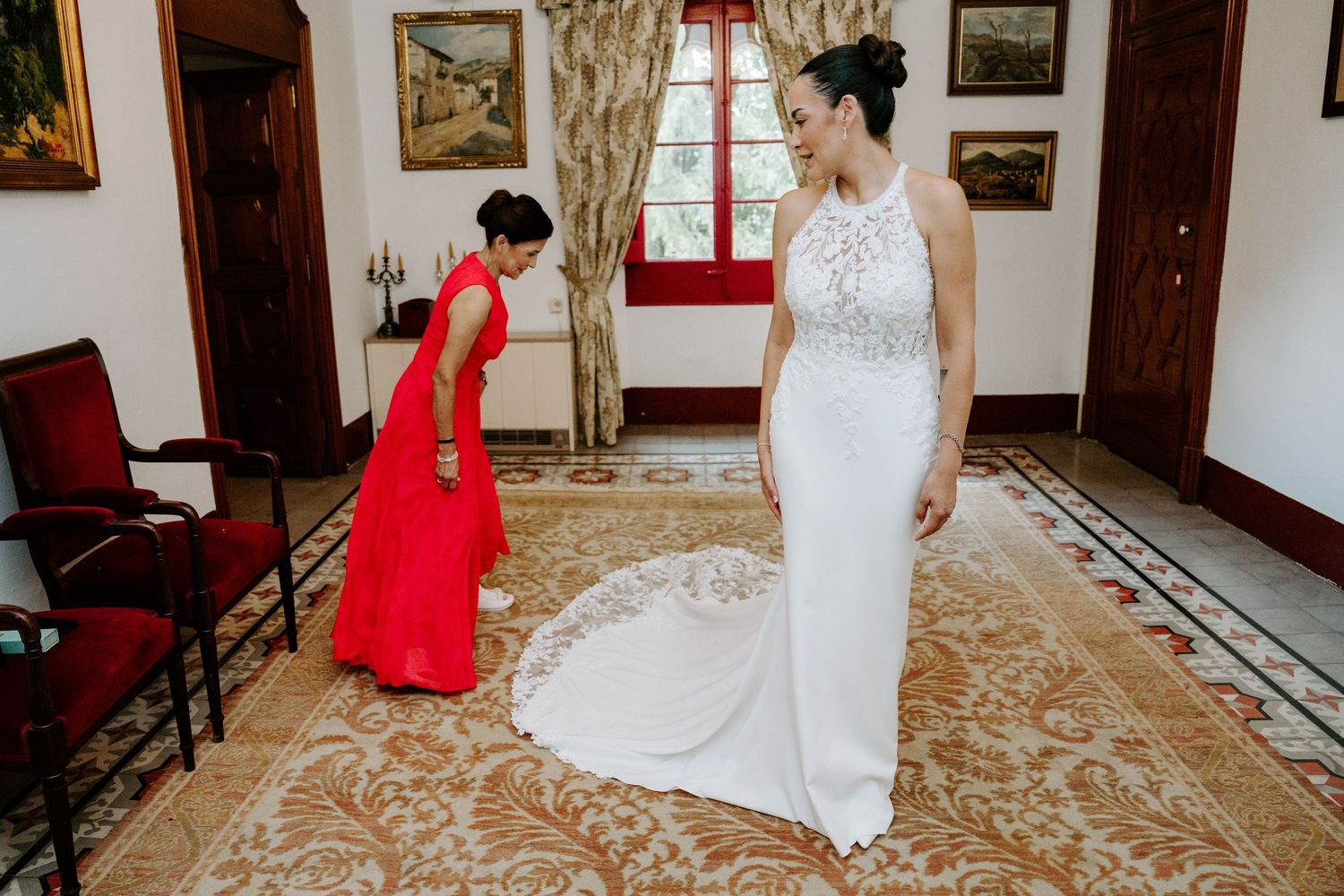 A bridesmaid in red adjusts the train of a bride's elegant white halter wedding gown in a classic interior.