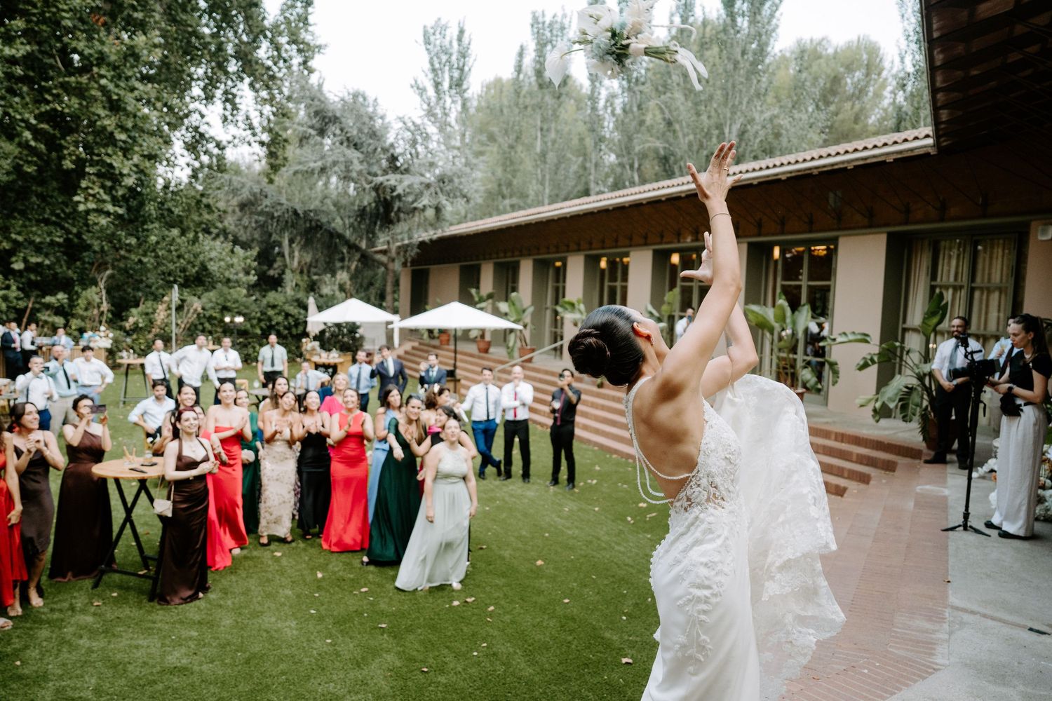 Bride tosses bouquet to excited wedding guests gathered on lush garden lawn during outdoor reception.