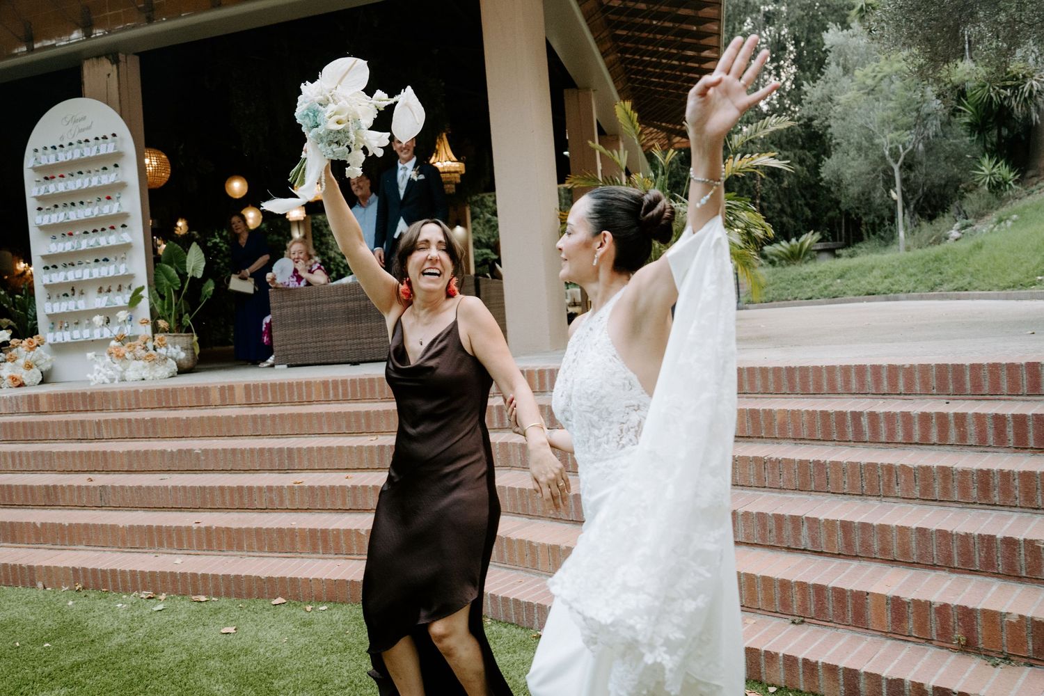 Bride in white gown and guest in black dress celebrate joyfully outdoors, raising hands with bouquet at wedding venue.