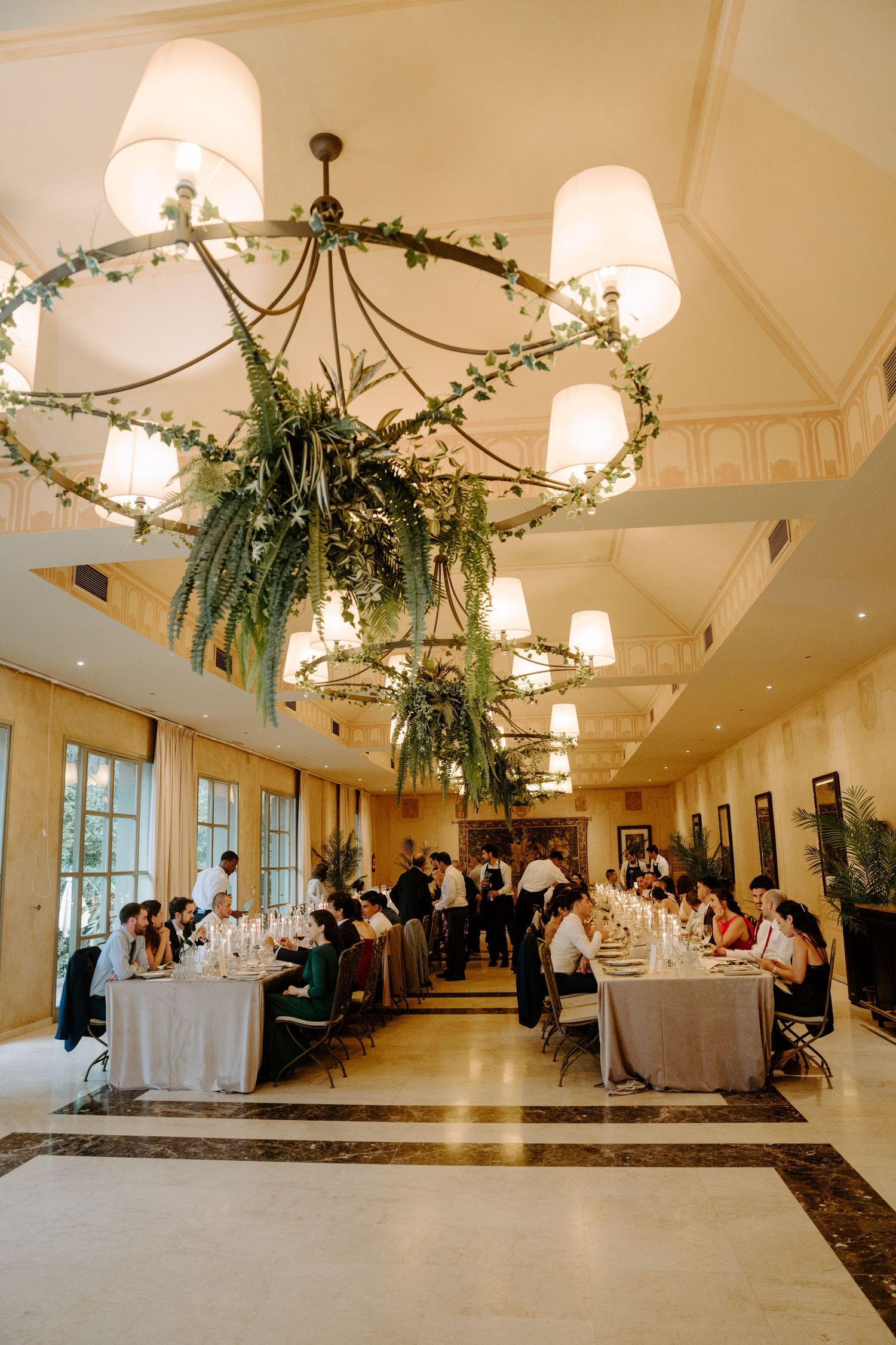 Elegant wedding reception hall with long dining tables, floral chandeliers, and guests seated beneath warm lighting.