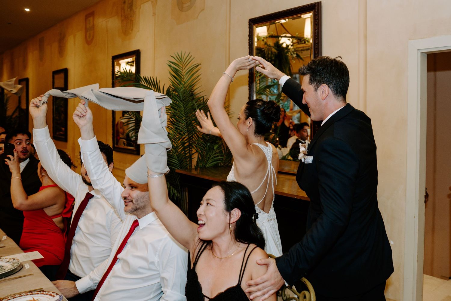 Wedding guests celebrate joyfully waving napkins at lively indoor wedding reception dinner.