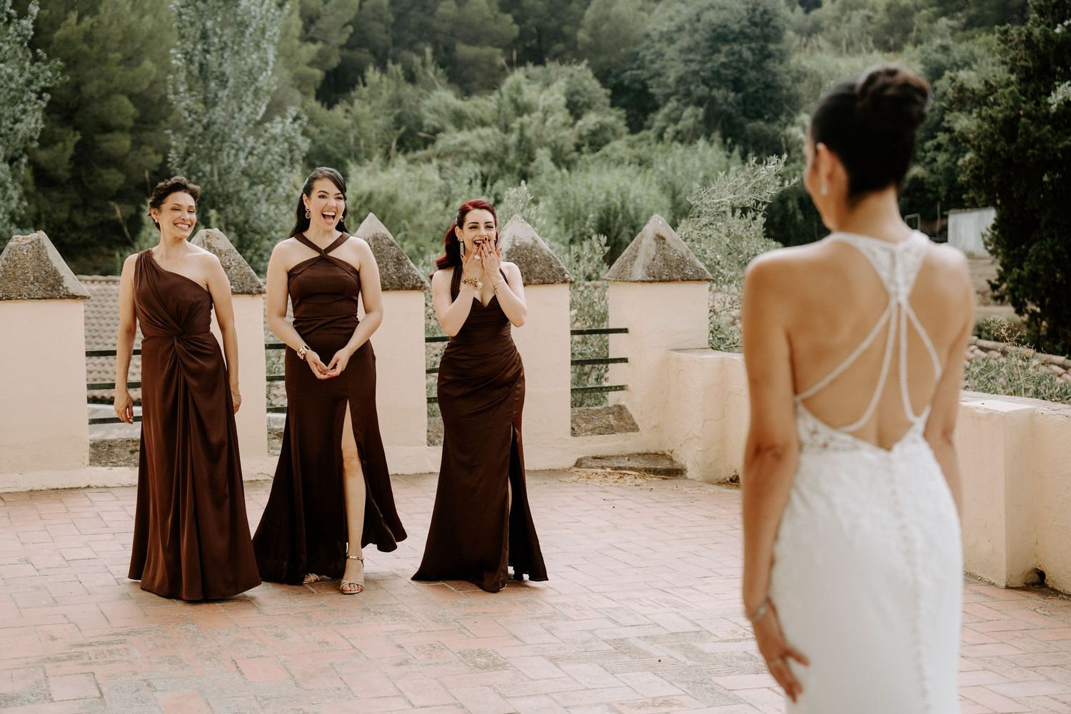 Three bridesmaids in brown gowns react with joy seeing bride in white lace halter wedding dress on terrace.