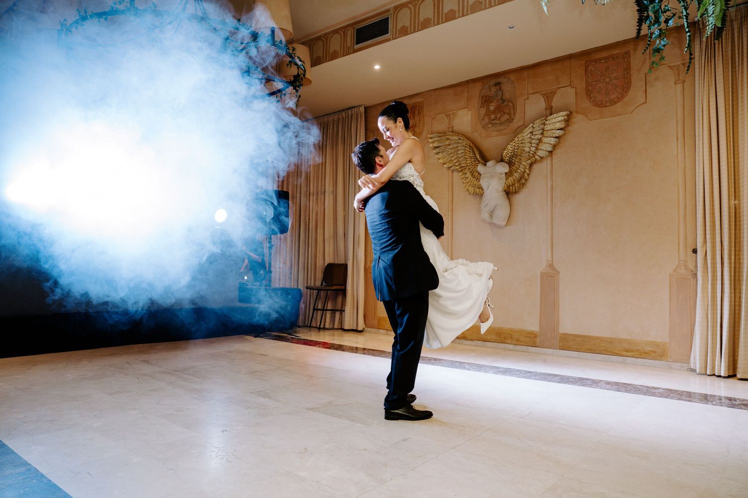 Groom lifts bride during romantic first dance surrounded by dramatic blue smoke on ballroom floor.