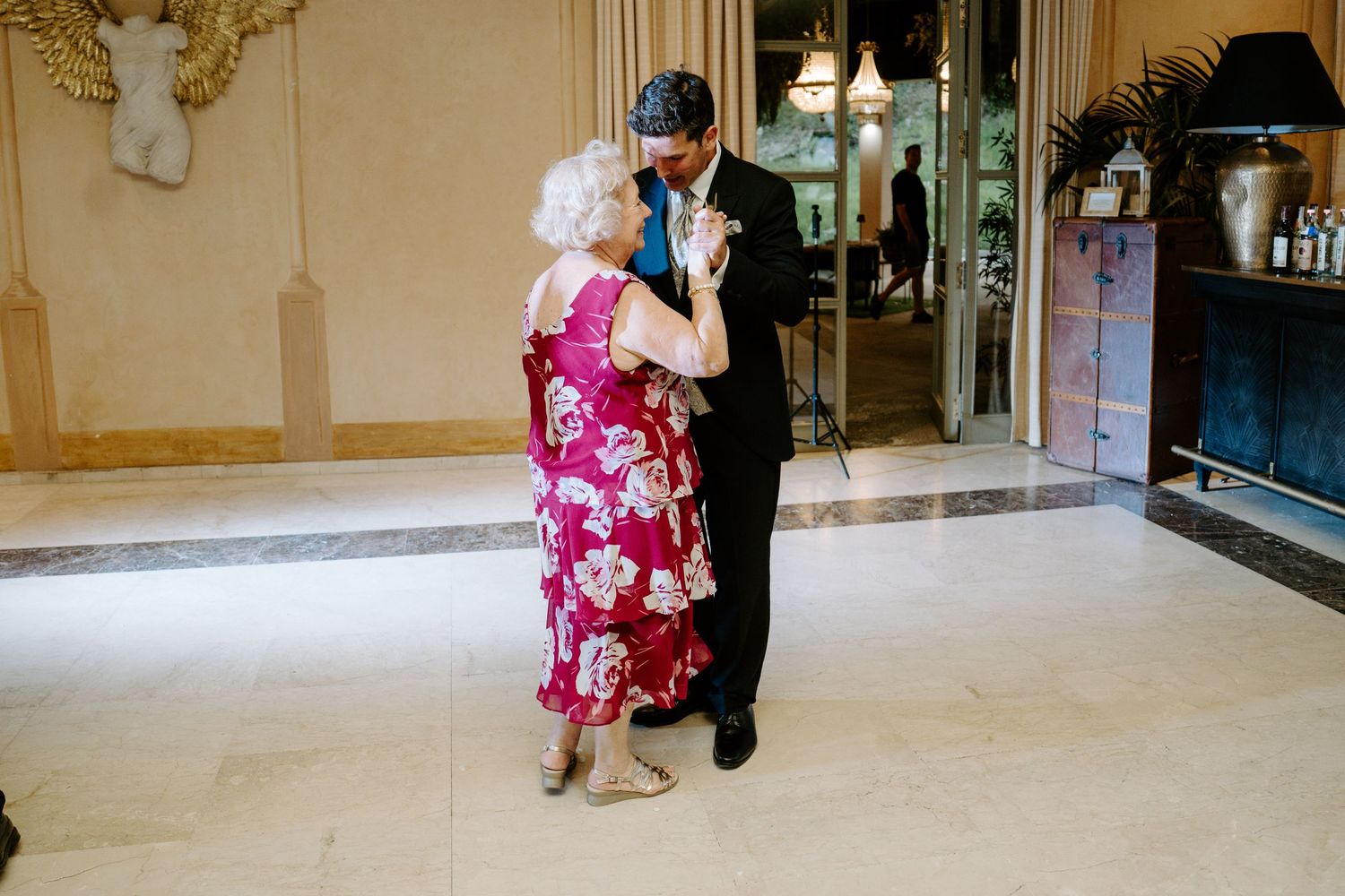 Elderly woman in floral dress dancing with a man in a suit at an elegant wedding reception venue.