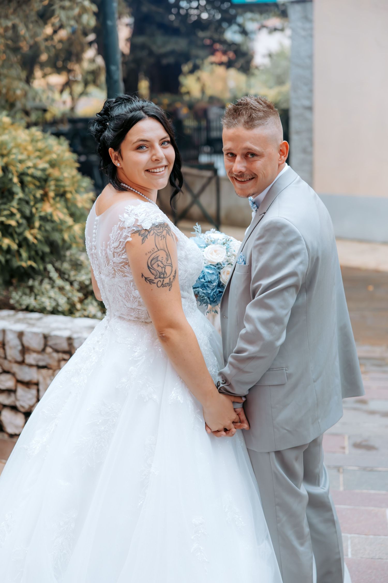 Bride and groom smiling at each other on a balcony, bride holding a white fan and blue floral bouquet.