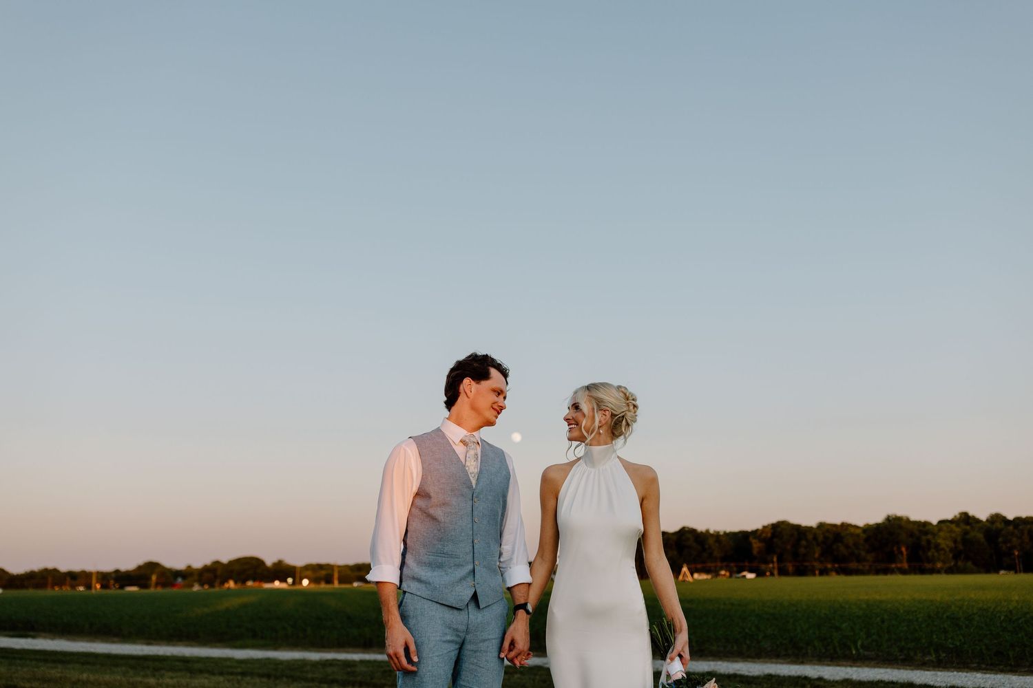 Bride and groom holding hands at sunset in an open field, wearing elegant wedding attire.