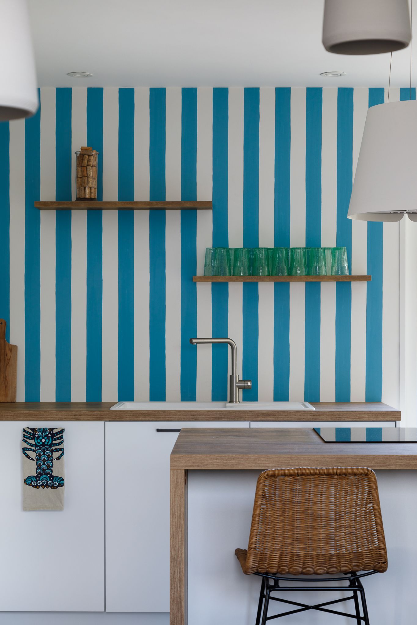Mur de cuisine aux rayures bleues et blanches audacieuses, avec des étagères en bois, de la verrerie verte et un tabouret de bar en rotin.