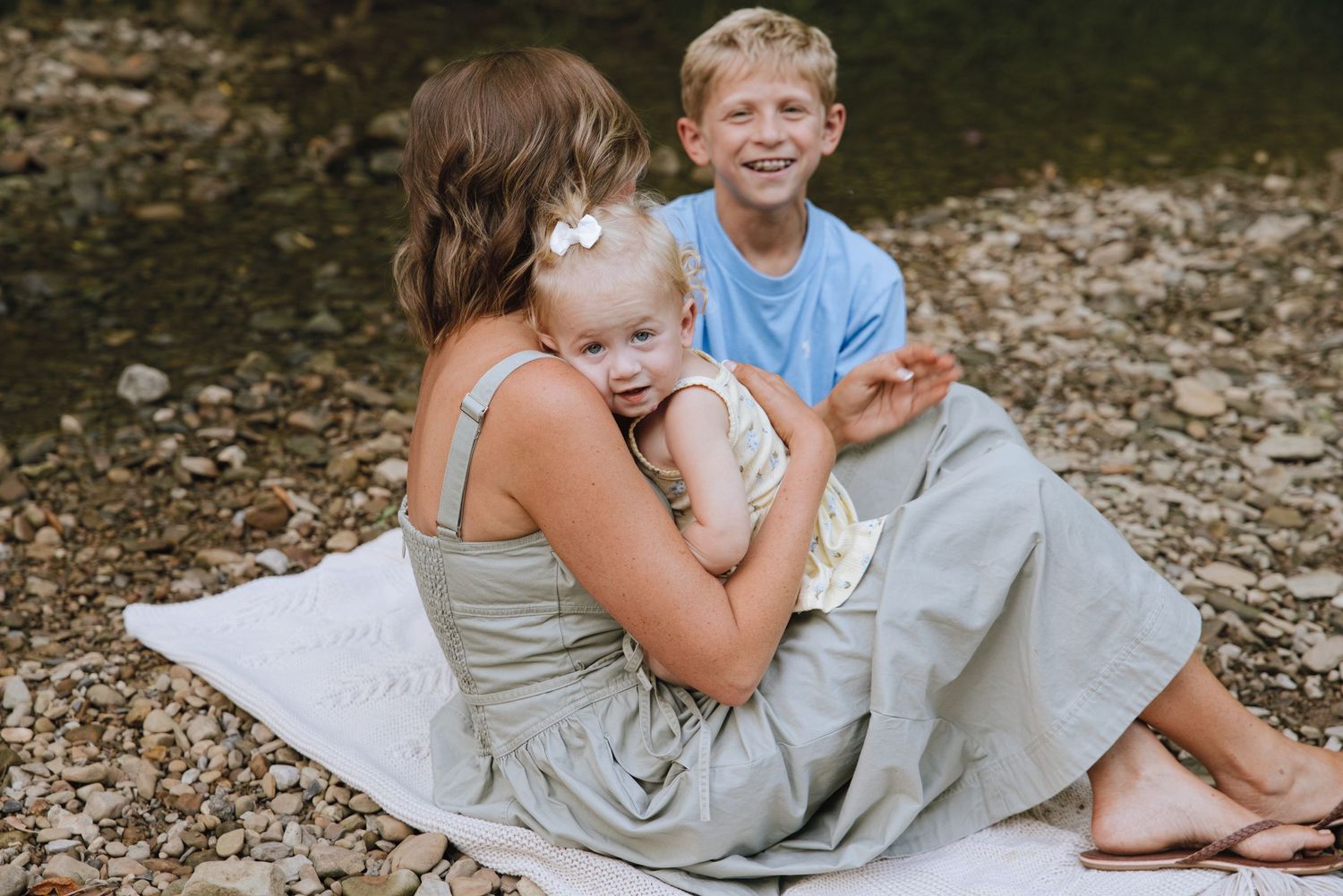 A mother sits on a blanket by a rocky creek, hugging her toddler while a smiling blonde boy sits beside them.