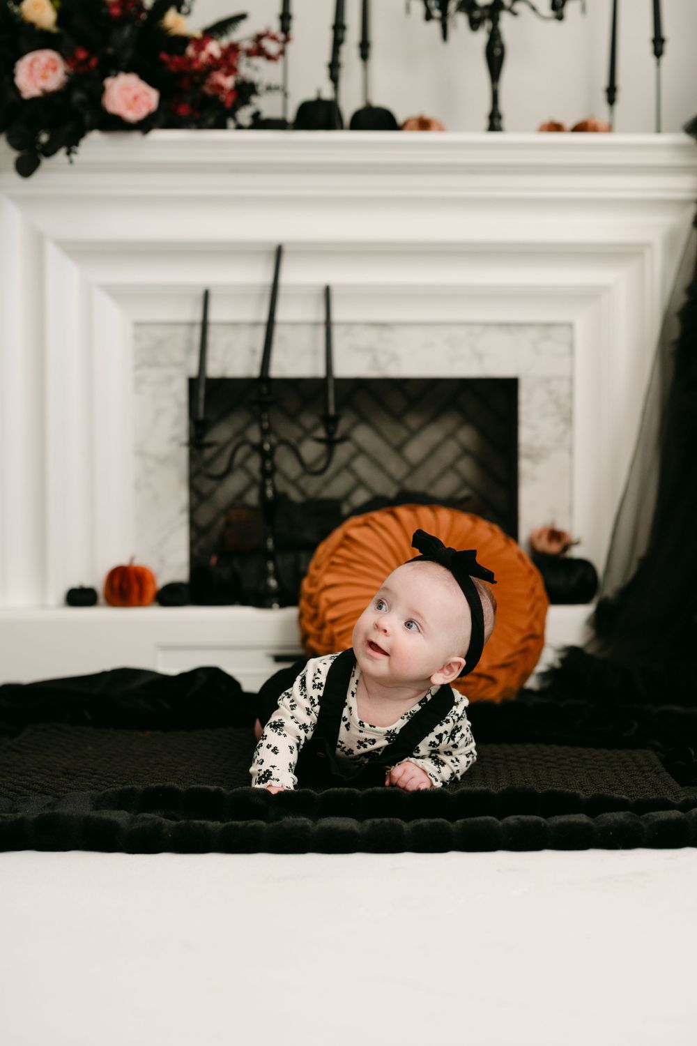 Baby in Halloween outfit doing tummy time on black blanket near decorated fireplace with pumpkins and spooky decor.
