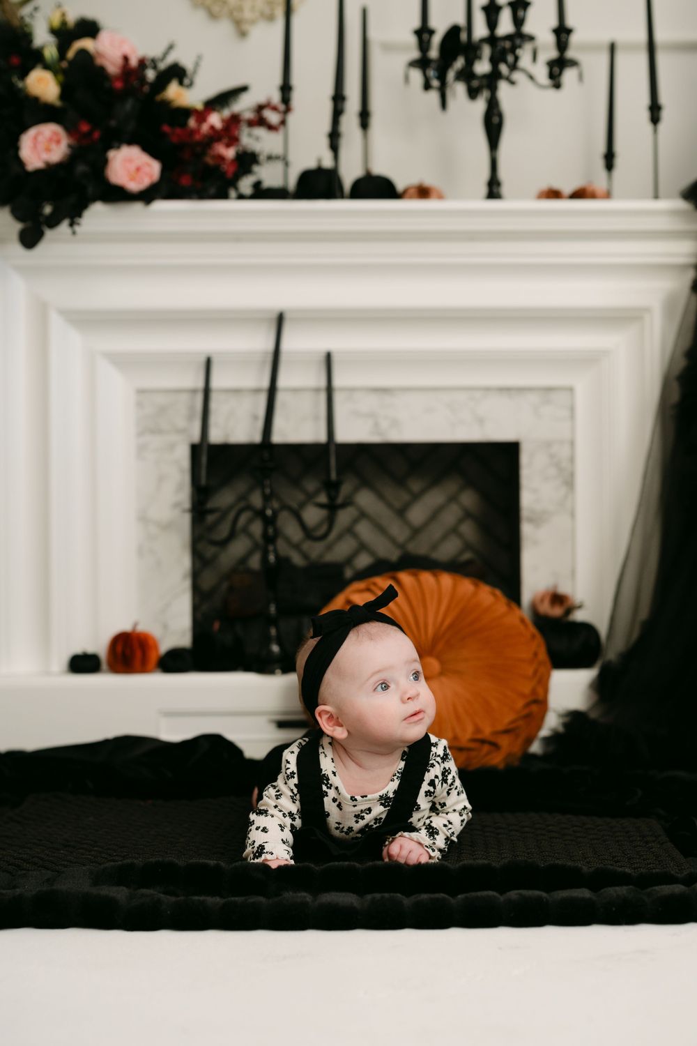 Baby in Halloween outfit lying on black blanket before decorated fireplace with pumpkins and dark florals.
