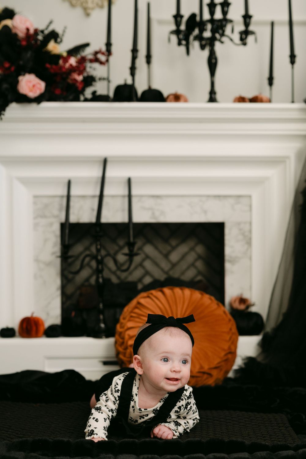 Baby in Halloween costume doing tummy time near decorated fireplace with pumpkins and black candelabra.