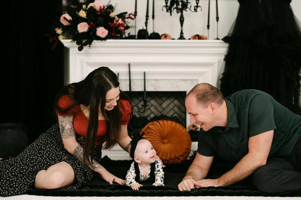 Family photo with parents smiling at their baby on a dark Halloween-themed set with floral decor and fireplace.