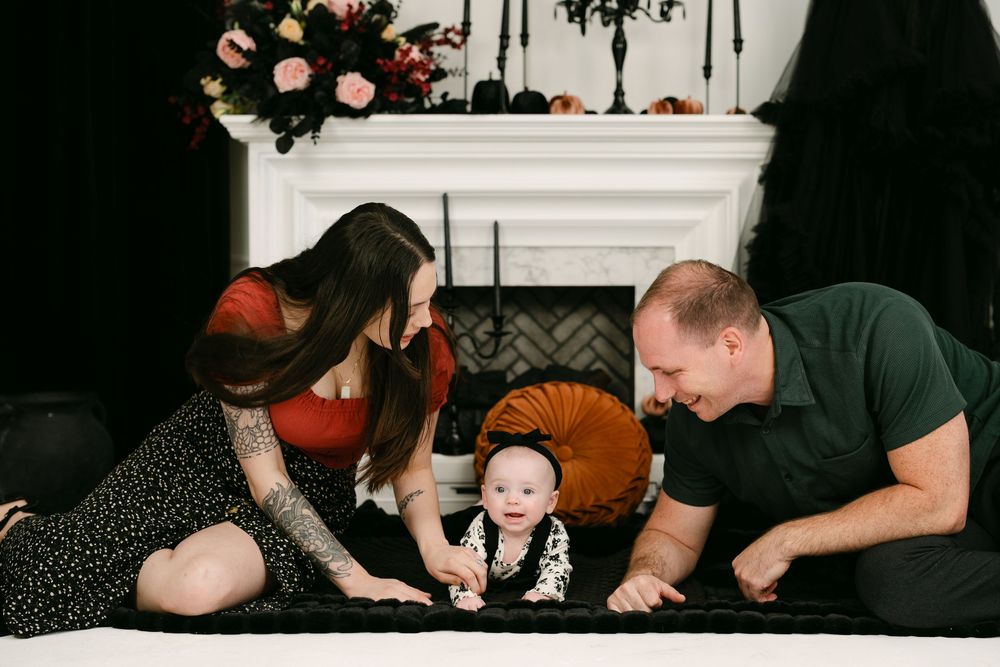 A young family with a tattooed mom and dad smiling at their baby on a blanket near a decorated fireplace.
