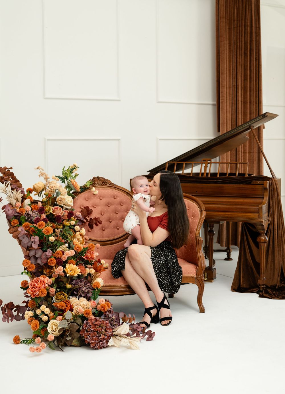 Mother kissing baby on pink velvet sofa beside autumn floral arrangement and grand piano in white studio.